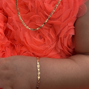 Puede incluir: Un conjunto de collar y pulsera de color dorado. Las joyas presentan un dise&ntilde;o de eslabones planos y rectangulares con detalles de estrella. Las piezas se exhiben sobre una tela naranja brillante y texturizada.