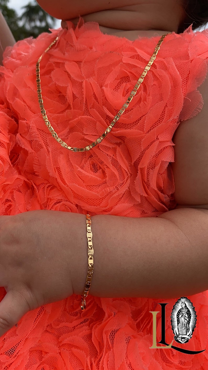 Puede incluir: Un conjunto de collar y pulsera de color dorado. Las joyas presentan un dise&ntilde;o de eslabones planos y rectangulares con detalles de estrella. Las piezas se exhiben sobre una tela naranja brillante y texturizada.