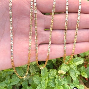 May include: A gold chain necklace with a delicate, patterned design. The chain is made of small, oval-shaped links with a textured surface. The necklace is shown on a hand, with the chain draped over the fingers.