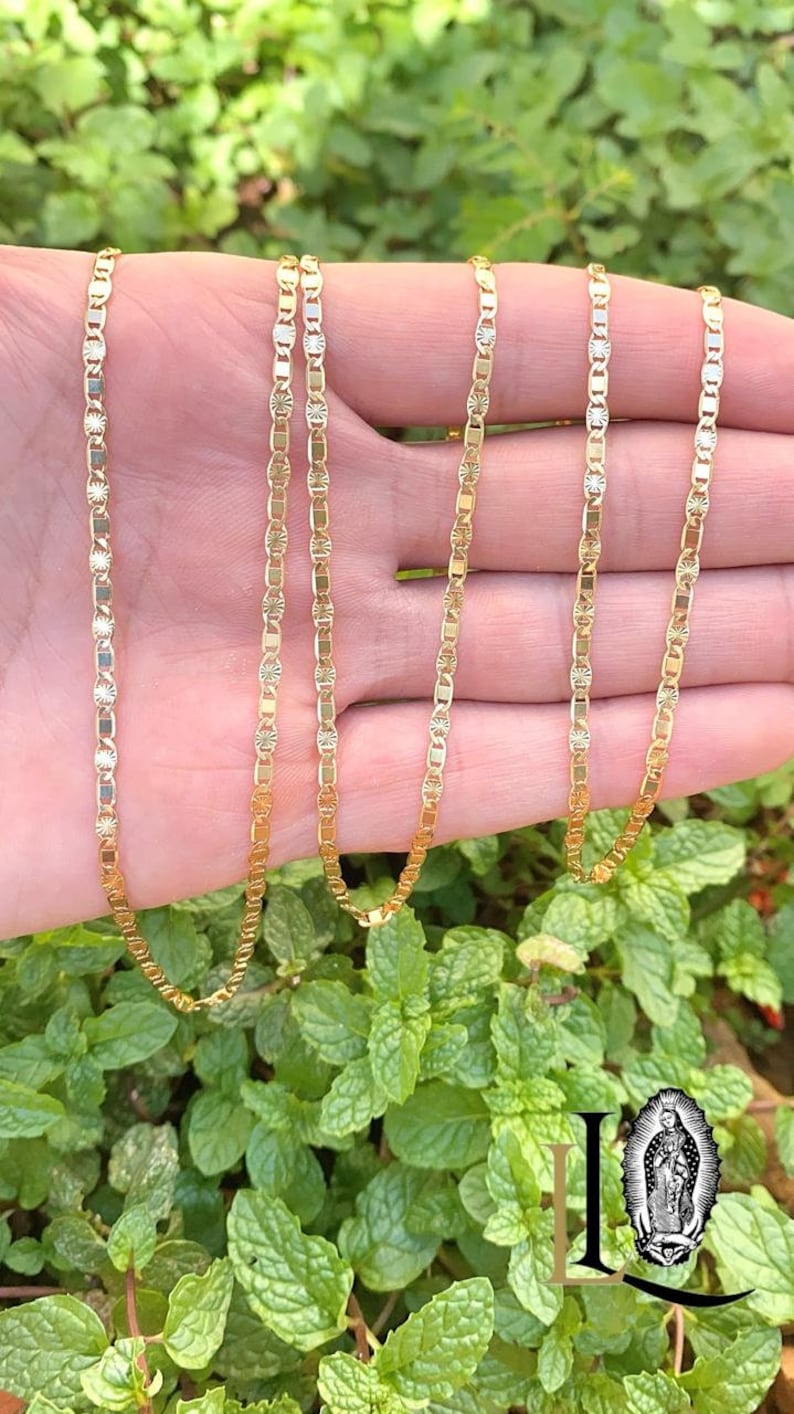 May include: A gold chain necklace with a delicate, patterned design. The chain is made of small, oval-shaped links with a textured surface. The necklace is shown on a hand, with the chain draped over the fingers.