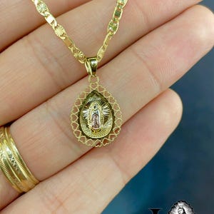 Puede incluir: Un collar dorado con un colgante que representa una figura religiosa. El colgante tiene forma de l&aacute;grima con un borde en forma de coraz&oacute;n. La cadena del collar tiene un dise&ntilde;o decorativo.