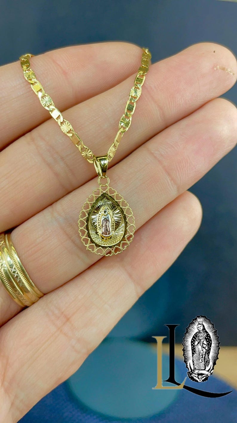 Puede incluir: Un collar dorado con un colgante que representa una figura religiosa. El colgante tiene forma de l&aacute;grima con un borde en forma de coraz&oacute;n. La cadena del collar tiene un dise&ntilde;o decorativo.