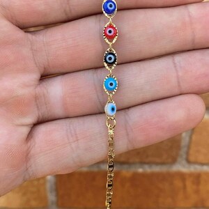 May include: A gold chain bracelet with five evil eye charms. The charms are blue, red, black, light blue, and white.