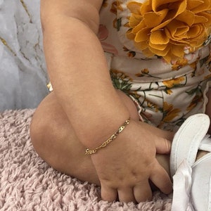 May include: A baby wearing a white shirt and floral overalls with a gold chain bracelet on their wrist.
