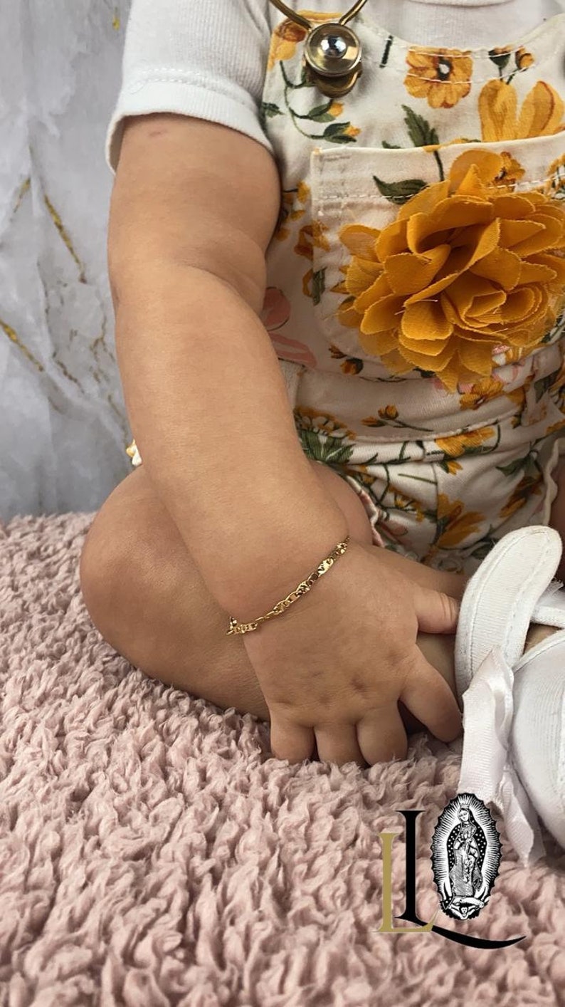 May include: A baby wearing a white shirt and floral overalls with a gold chain bracelet on their wrist.