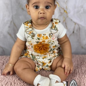 May include: A baby girl wearing a white onesie with a floral print and a yellow flower on the front. She is wearing a pink headband and white shoes.