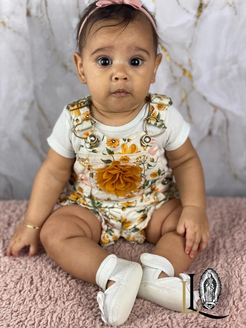 May include: A baby girl wearing a white onesie with a floral print and a yellow flower on the front. She is wearing a pink headband and white shoes.