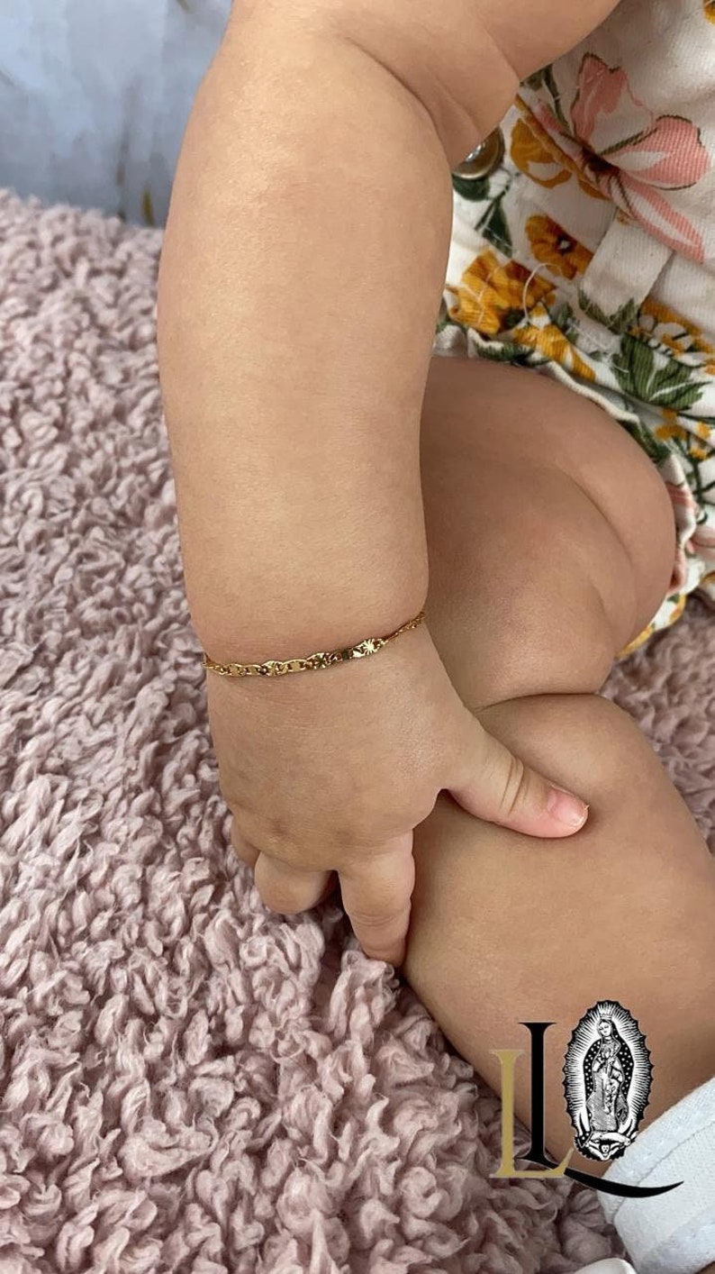 May include: A gold chain bracelet on a baby's wrist. The bracelet is a delicate, thin chain with a small, decorative charm.