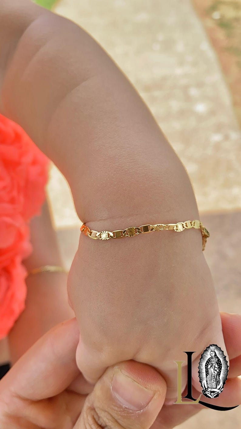 May include: A gold chain bracelet with a delicate, patterned design. The bracelet is worn on a child's wrist.