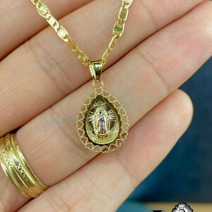 Puede incluir: Collar dorado con un colgante religioso. El colgante tiene forma de l&aacute;grima, con un borde en forma de coraz&oacute;n. La cadena tiene un patr&oacute;n decorativo. Tambi&eacute;n se ve un anillo dorado. Joyer&iacute;a religiosa.