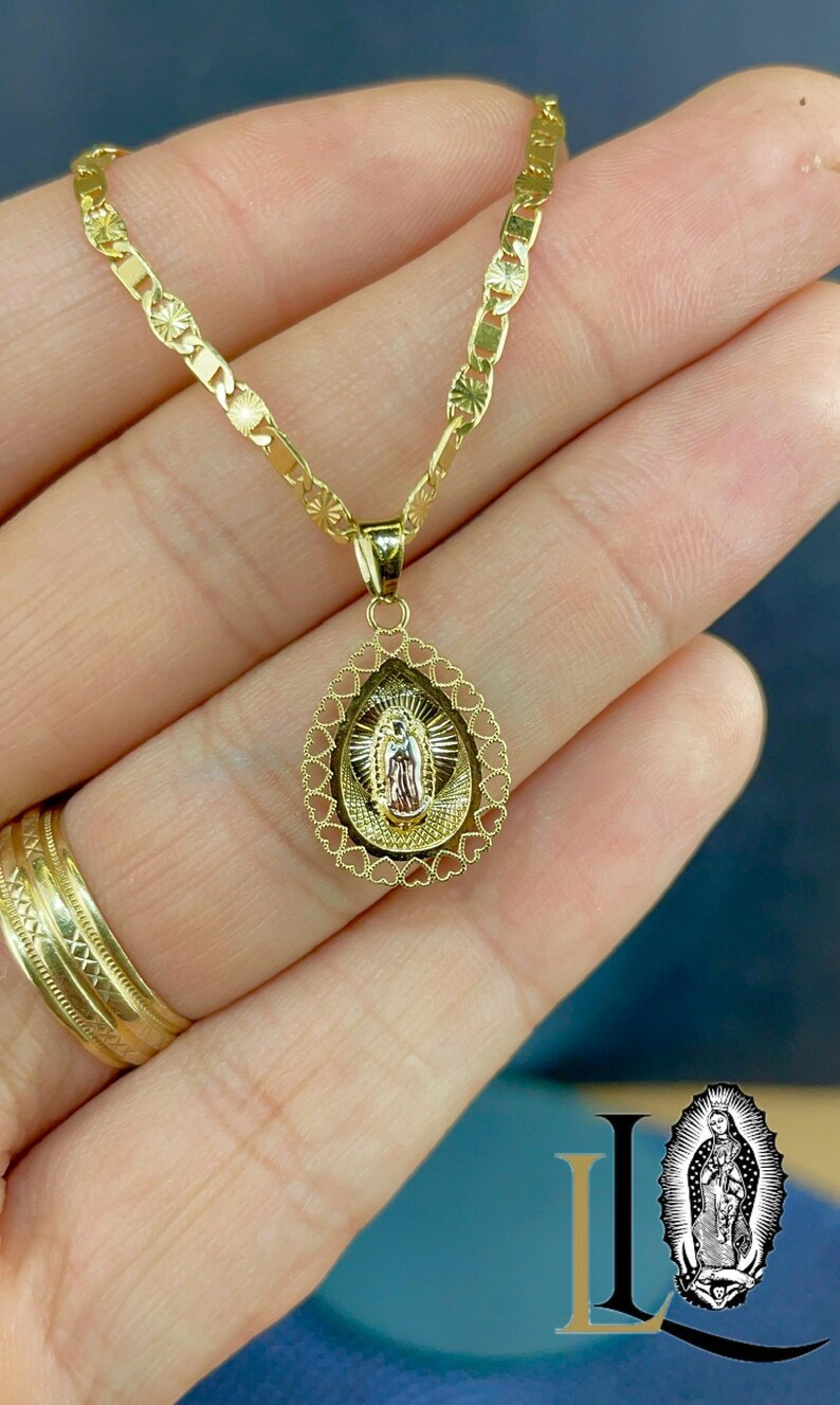 Puede incluir: Collar dorado con un colgante religioso. El colgante tiene forma de l&aacute;grima, con un borde en forma de coraz&oacute;n. La cadena tiene un patr&oacute;n decorativo. Tambi&eacute;n se ve un anillo dorado. Joyer&iacute;a religiosa.