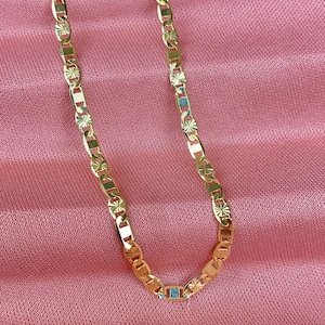 May include: Gold-colored chain necklace with a rectangular link design and starburst accents. The necklace is displayed on a pink fabric background. A small, light blue rectangular stone is set in the chain.