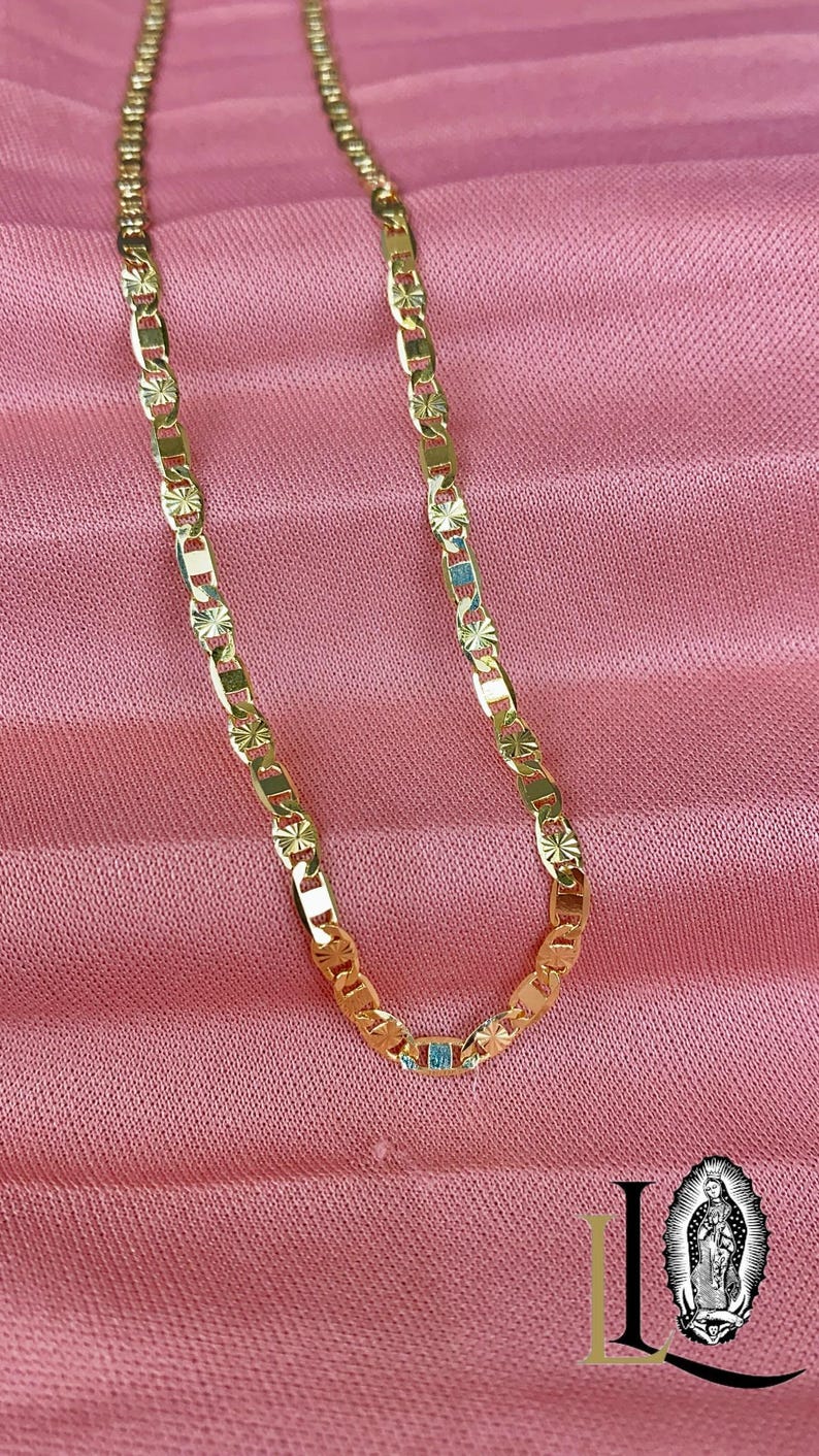 May include: Gold-colored chain necklace with a rectangular link design and starburst accents. The necklace is displayed on a pink fabric background. A small, light blue rectangular stone is set in the chain.