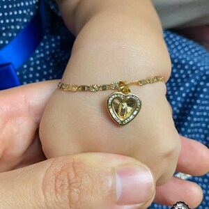 May include: A gold bracelet with a heart-shaped pendant. The pendant features a religious figure and is decorated with small, clear stones. The bracelet is on a child's wrist, held by an adult's hand.