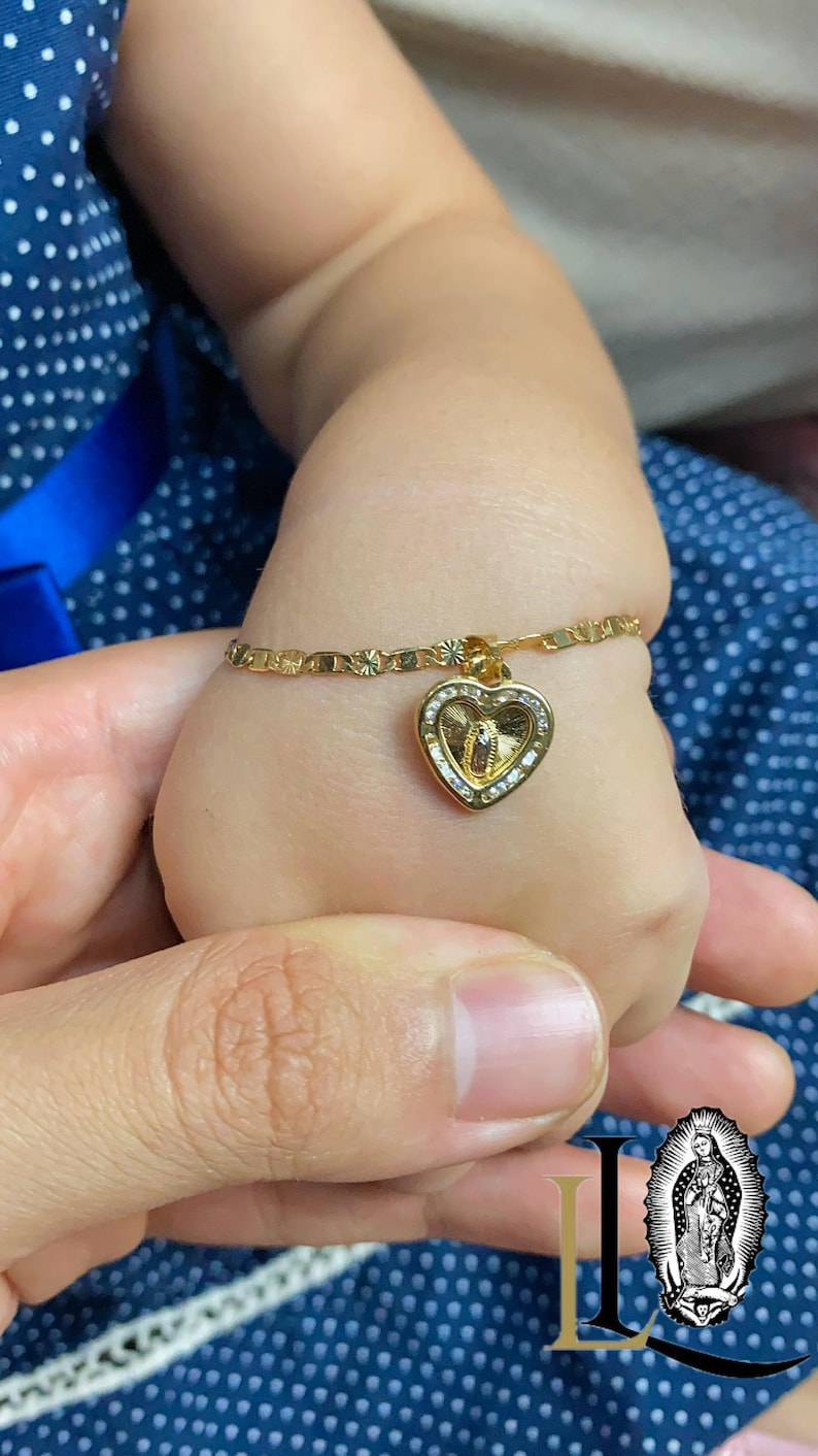 May include: A gold bracelet with a heart-shaped pendant. The pendant features a religious figure and is decorated with small, clear stones. The bracelet is on a child's wrist, held by an adult's hand.