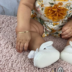 May include: A baby wearing a white floral romper with a yellow flower accent and white shoes. The baby is wearing a gold bracelet on their wrist.