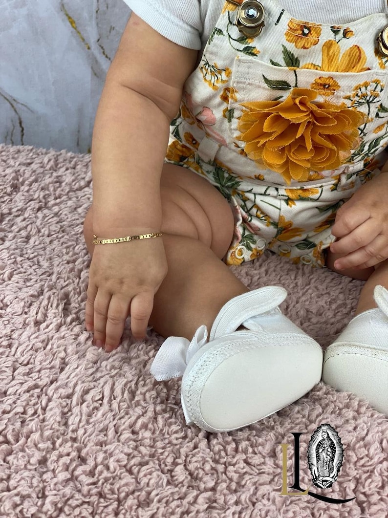 May include: A baby wearing a white floral romper with a yellow flower accent and white shoes. The baby is wearing a gold bracelet on their wrist.