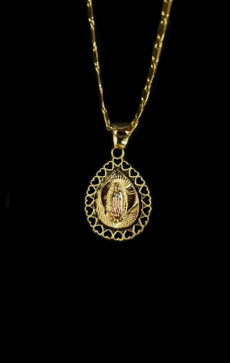 Puede incluir: Collar con colgante dorado que presenta un icono religioso dentro de un marco en forma de l&aacute;grima. El marco est&aacute; adornado con recortes en forma de coraz&oacute;n. La cadena y el colgante son de metal brillante y dorado.