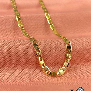 May include: A gold-colored chain necklace with a repeating pattern of rectangular and oval links. The links have a starburst design. The necklace is displayed on a pink fabric background.