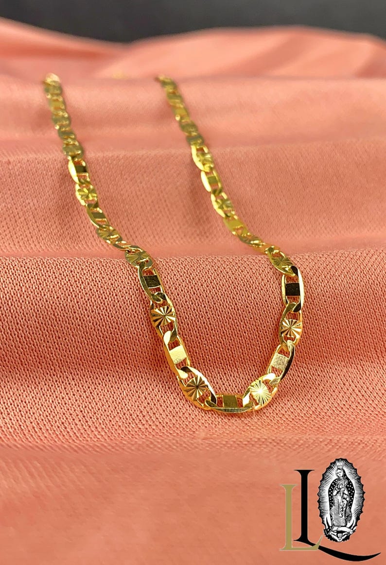 May include: A gold-colored chain necklace with a repeating pattern of rectangular and oval links. The links have a starburst design. The necklace is displayed on a pink fabric background.