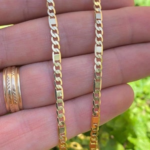 May include: Two gold chains, one thicker than the other, are shown on a hand. The chains are both made of a flat, rectangular link design.