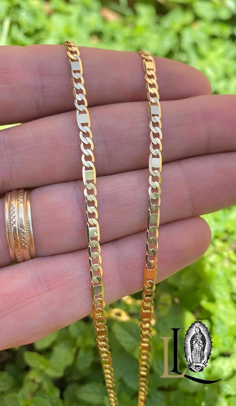 May include: Two gold chains, one thicker than the other, are shown on a hand. The chains are both made of a flat, rectangular link design.