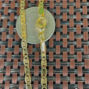 May include: A gold-colored chain necklace with a decorative pattern. The chain is shown next to a quarter for size comparison. The necklace has a lobster clasp closure. The background is a brown and black woven pattern.