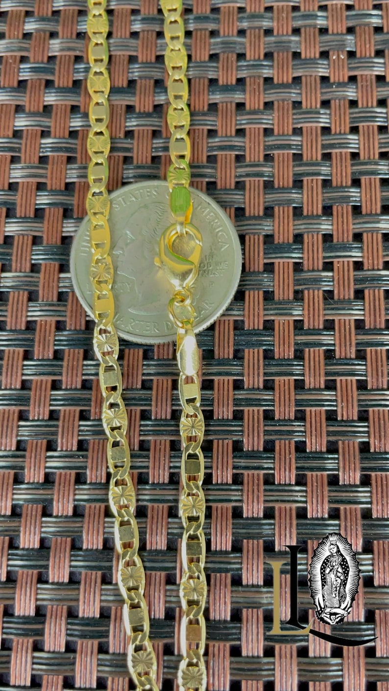 May include: A gold-colored chain necklace with a decorative pattern. The chain is shown next to a quarter for size comparison. The necklace has a lobster clasp closure. The background is a brown and black woven pattern.