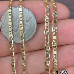 May include: Close-up of three gold-colored chain necklaces with a decorative pattern. A US quarter is next to the chains for size comparison. The chains have a repeating pattern of rectangular and star-shaped links. The chains are displayed on a brown surface.