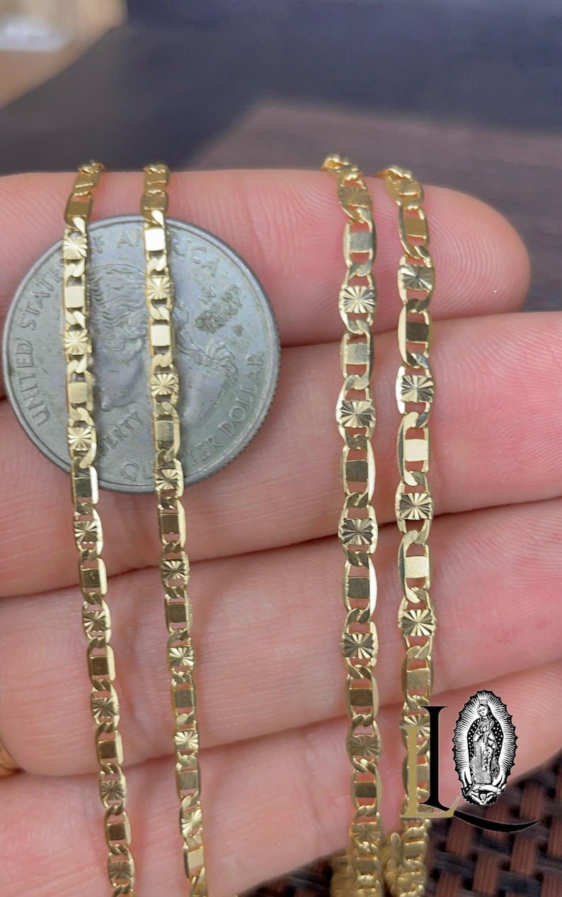 May include: Close-up of three gold-colored chain necklaces with a decorative pattern. A US quarter is next to the chains for size comparison. The chains have a repeating pattern of rectangular and star-shaped links. The chains are displayed on a brown surface.