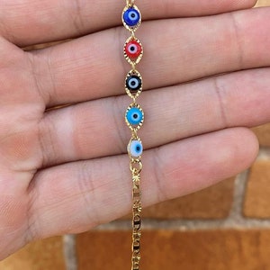 May include: Gold chain bracelet with five evil eye charms. The charms are blue, red, black, light blue, and white.