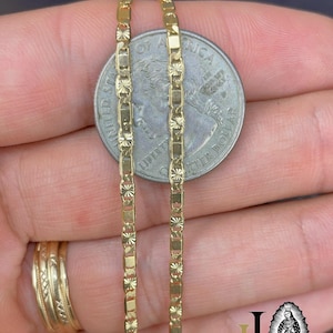 May include: Close-up of two gold-colored chain necklaces with a quarter coin for size comparison. The chains have a rectangular and floral design. A gold ring is visible on the left. The image is well-lit, highlighting the jewelry's details.