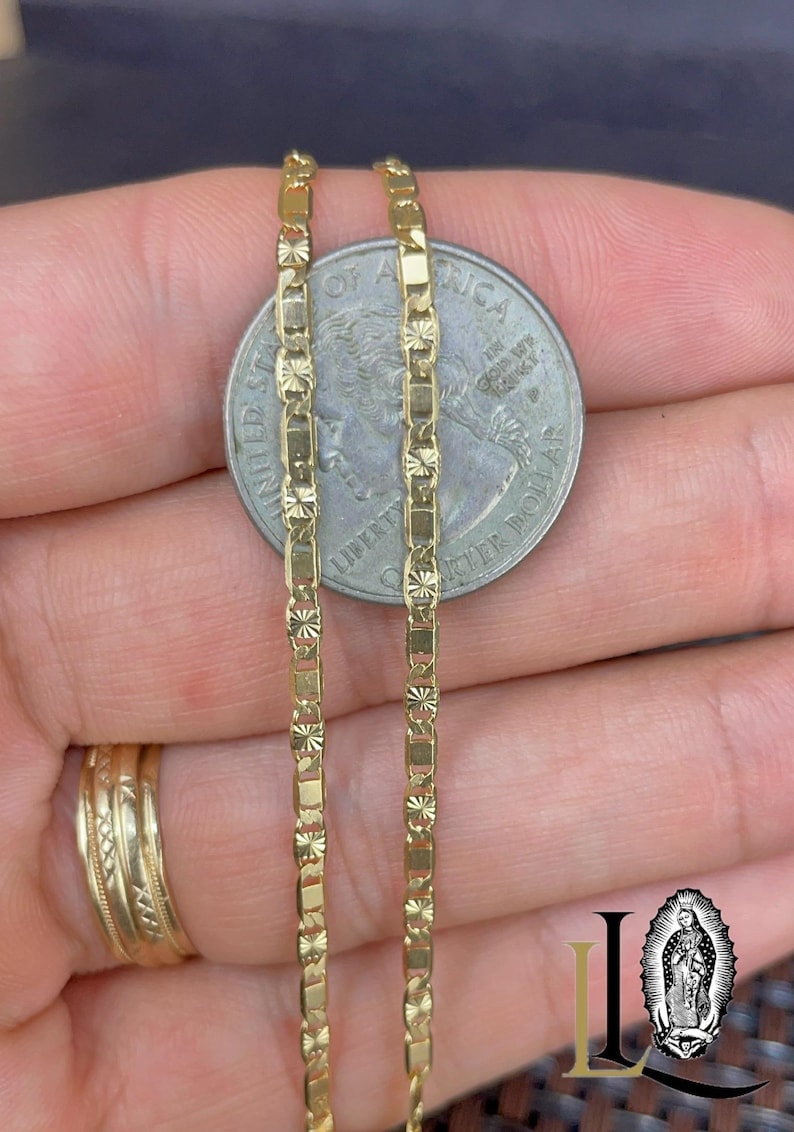 May include: Close-up of two gold-colored chain necklaces with a quarter coin for size comparison. The chains have a rectangular and floral design. A gold ring is visible on the left. The image is well-lit, highlighting the jewelry's details.