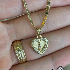 May include: A gold necklace with a heart-shaped pendant featuring a religious figure. The pendant is adorned with small, clear stones and a detailed image. A matching gold ring with an intricate design is also visible.