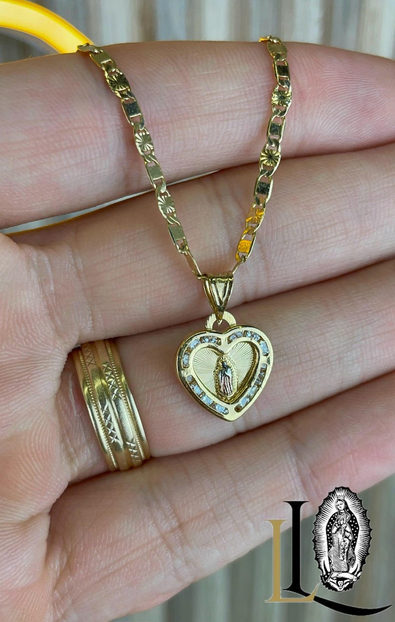 May include: A gold necklace with a heart-shaped pendant featuring a religious figure. The pendant is adorned with small, clear stones and a detailed image. A matching gold ring with an intricate design is also visible.