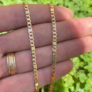 May include: Two gold chains, one on top of the other, are held in a hand. The chains are made of a flat, rectangular link design.