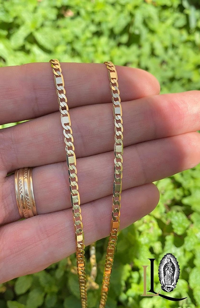 May include: Two gold chains, one on top of the other, are held in a hand. The chains are made of a flat, rectangular link design.