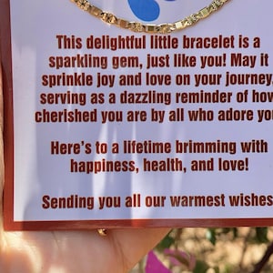 May include: A gold chain bracelet with a delicate design. The bracelet is displayed on a card with a message about joy, love, and happiness. The card also features a pair of blue footprints.