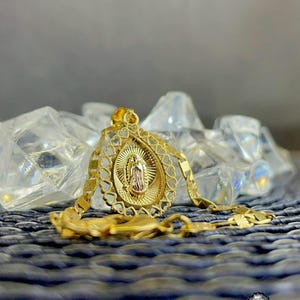 Puede incluir: Un collar con colgante dorado que presenta una representaci&oacute;n detallada de una figura religiosa dentro de un marco ovalado. El colgante cuelga de una cadena a juego, descansando sobre una superficie texturizada. La imagen incluye un logotipo en la esquina inferior derecha.