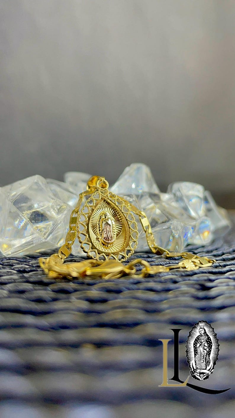 Puede incluir: Un collar con colgante dorado que presenta una representaci&oacute;n detallada de una figura religiosa dentro de un marco ovalado. El colgante cuelga de una cadena a juego, descansando sobre una superficie texturizada. La imagen incluye un logotipo en la esquina inferior derecha.