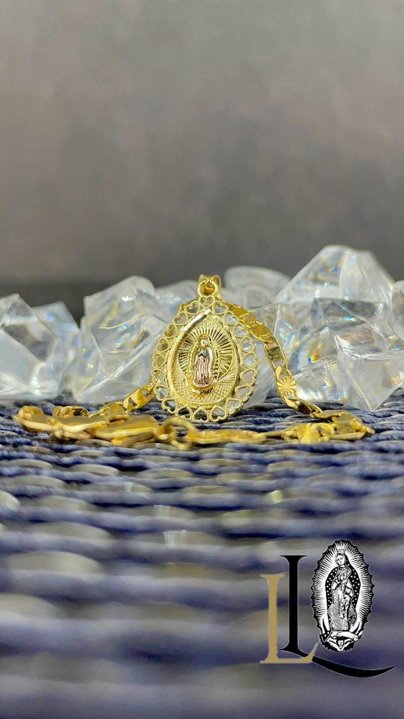 Puede incluir: Un collar con colgante dorado con un icono religioso dentro de un marco ovalado. El colgante cuelga de una cadena a juego. El fondo incluye cristales claros y facetados y una superficie azul texturizada.