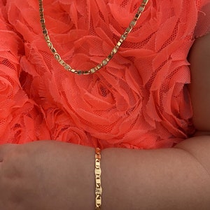 Puede incluir: Un conjunto de collar y pulsera de color dorado. El collar est&aacute; alrededor del cuello y la pulsera en la mu&ntilde;eca. Las joyas est&aacute;n en una persona que lleva un vestido color coral. Las joyas tienen un patr&oacute;n repetitivo de eslabones rectangulares con detalles de estrella.