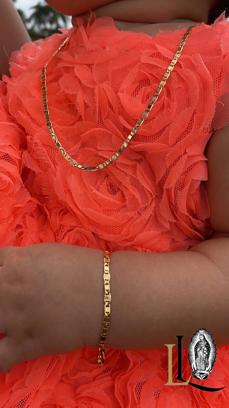 Puede incluir: Un conjunto de collar y pulsera de color dorado. El collar est&aacute; alrededor del cuello y la pulsera en la mu&ntilde;eca. Las joyas est&aacute;n en una persona que lleva un vestido color coral. Las joyas tienen un patr&oacute;n repetitivo de eslabones rectangulares con detalles de estrella.