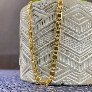 May include: A gold-colored chain necklace with a repeating pattern of rectangular and starburst-shaped links. The necklace is displayed against a textured, light gray background with a green plant and a blue surface.