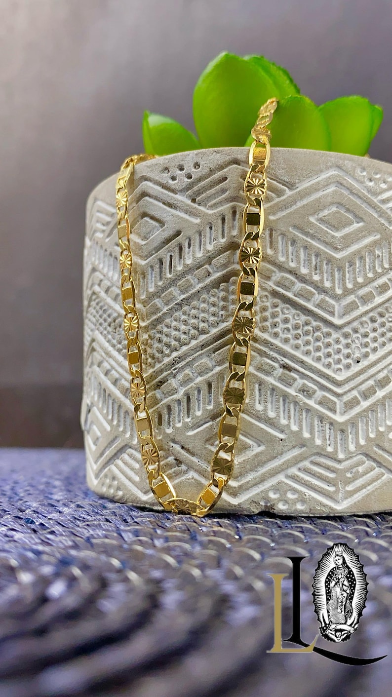 May include: A gold-colored chain necklace with a repeating pattern of rectangular and starburst-shaped links. The necklace is displayed against a textured, light gray background with a green plant and a blue surface.