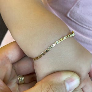 May include: A gold chain bracelet with a delicate design. The bracelet is on a baby's wrist.
