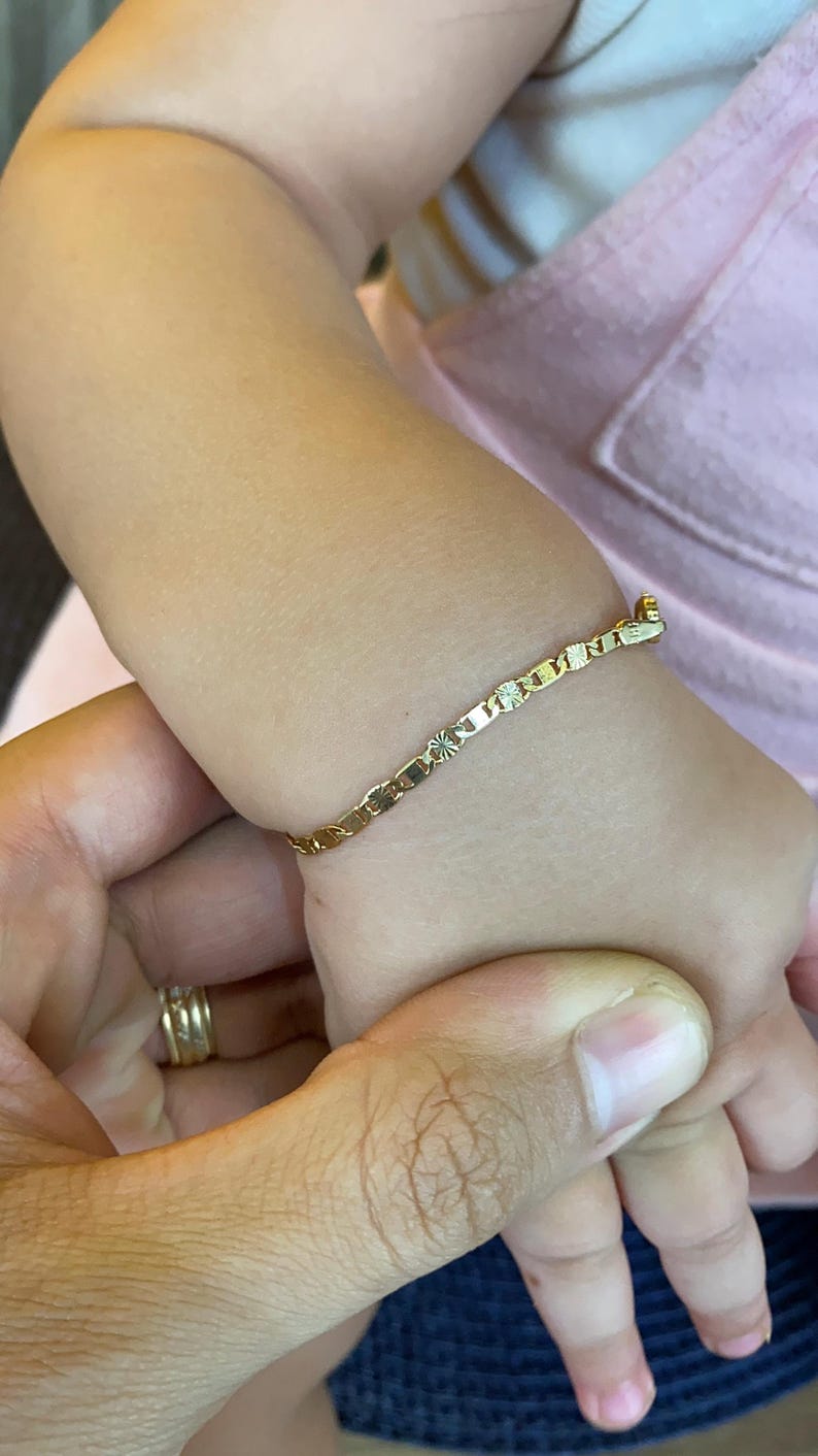 May include: A gold chain bracelet with a delicate design. The bracelet is on a baby's wrist.