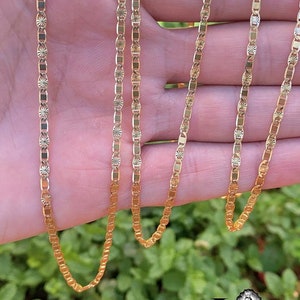 May include: A gold chain necklace with a delicate, patterned design. The chain is made of multiple strands and is shown draped over a hand.