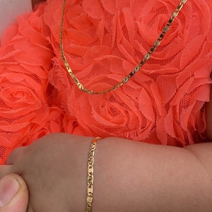 Puede incluir: Un conjunto de collar y pulsera de color dorado. Las joyas est&aacute;n en el brazo de un ni&ntilde;o, sobre un vestido naranja brillante. La pulsera y el collar tienen un patr&oacute;n repetido de peque&ntilde;as formas decorativas. Una mano con un anillo de oro sostiene el brazo del ni&ntilde;o.