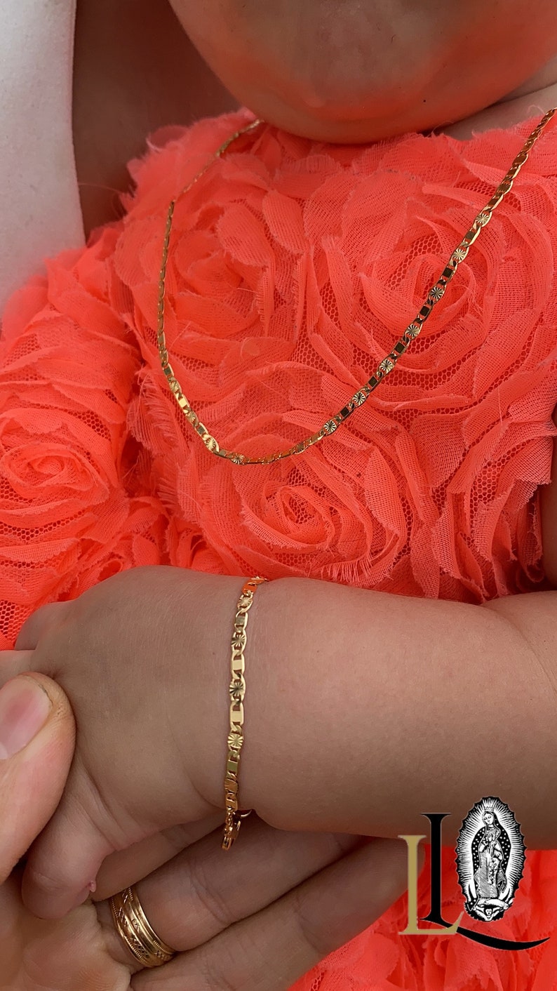Puede incluir: Un conjunto de collar y pulsera de color dorado. Las joyas est&aacute;n en el brazo de un ni&ntilde;o, sobre un vestido naranja brillante. La pulsera y el collar tienen un patr&oacute;n repetido de peque&ntilde;as formas decorativas. Una mano con un anillo de oro sostiene el brazo del ni&ntilde;o.
